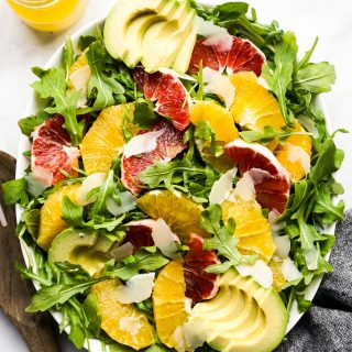 An oval bowl of arugula topped with oranges, avocados and parmesan cheese