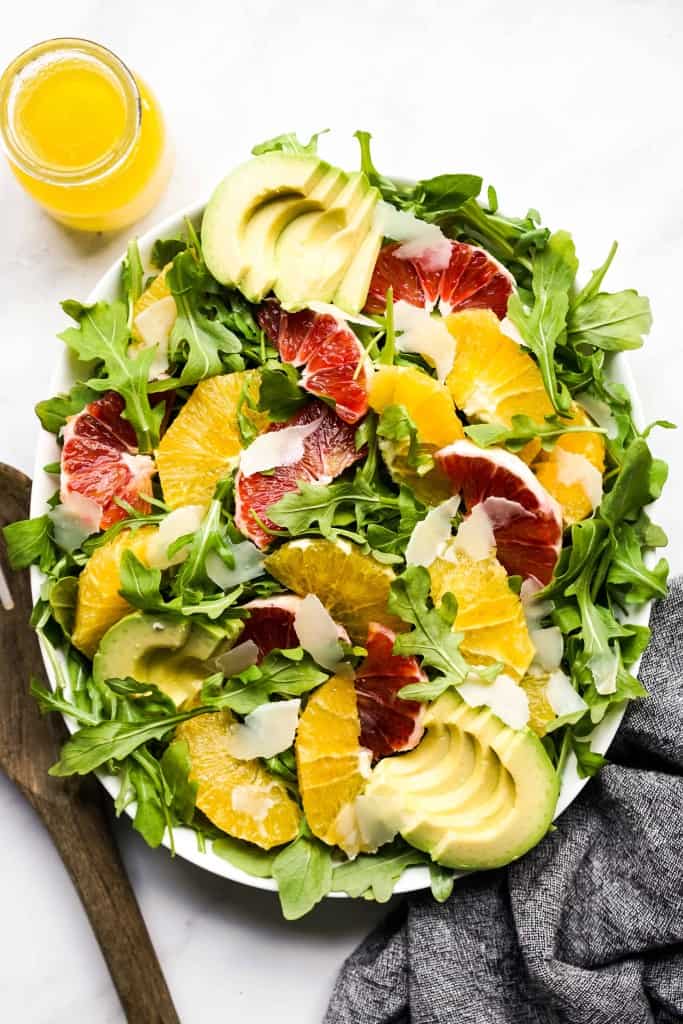 An oval bowl of arugula topped with oranges, avocados and parmesan cheese