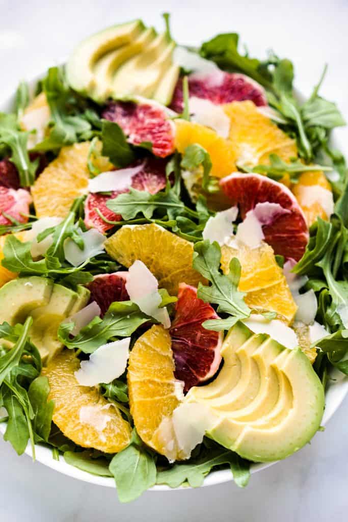 closeup of a bowl of arugula topped with oranges, avocados and parmesan