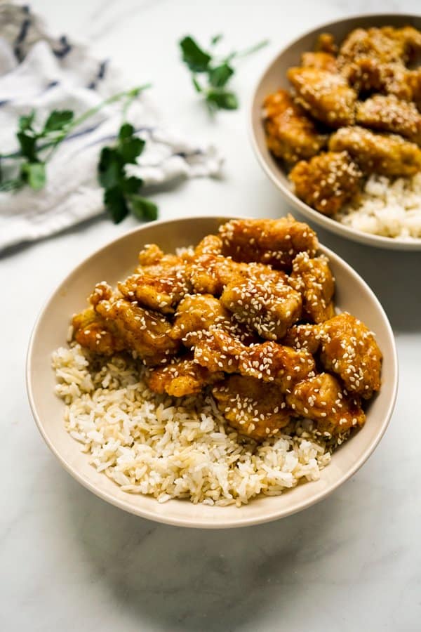 A bowl of crispy sesame chicken on top of rice with another bowl in the background