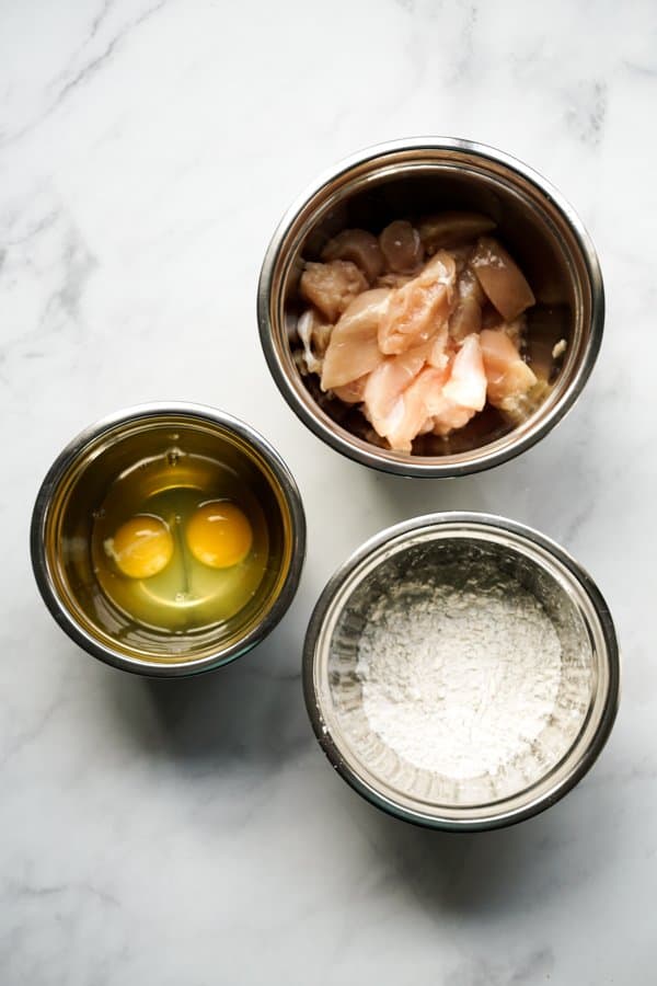 A bowl of raw chicken, a nowl of flour mixture and a bowl of two raw eggs