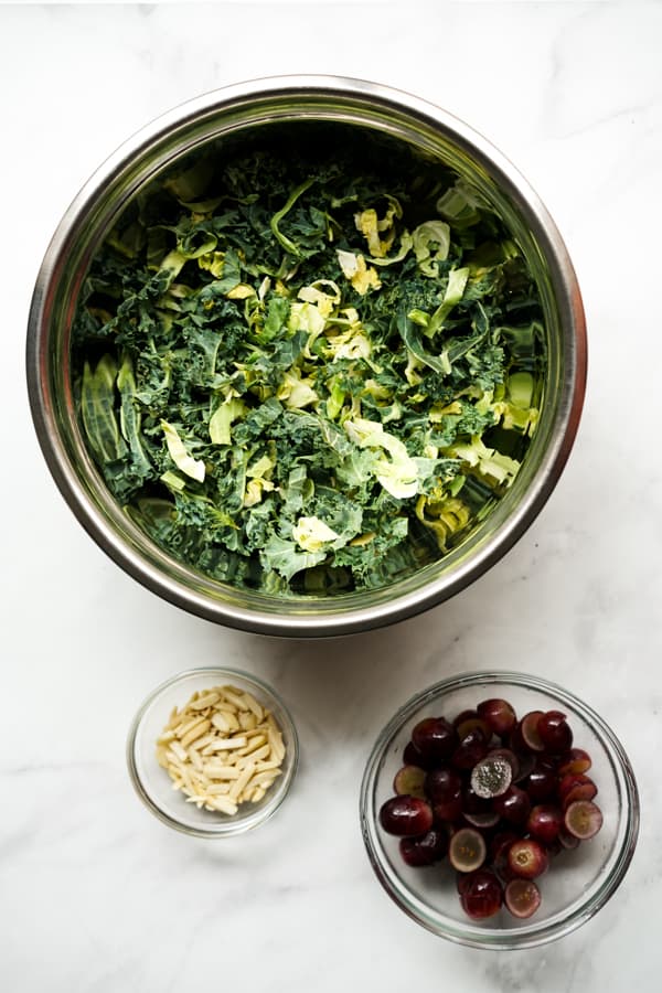 A bowl of chopped kale and Brussels sprouts, a bowl of grape, and almonds