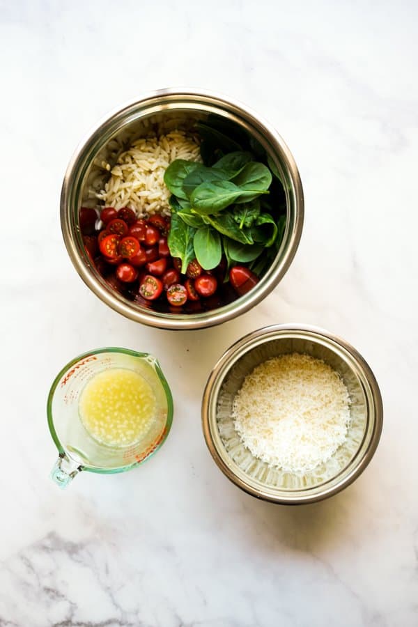 A bowl of pasta with cherry tomatoes and spinach, a bowl of shredded parmesan cheese, and a cup of lemon juice