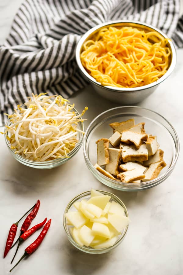 A bowl of yellow noodles, a bowl of beans sprouts, fried tofu, sliced potatoes and red chilies