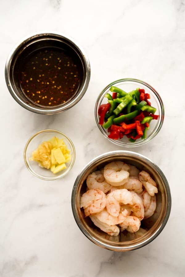 A bowl of raw shrimp, a bown of garlic and ginger, a bowl of red and green bell peppers, and a bowl of General Tso sauce