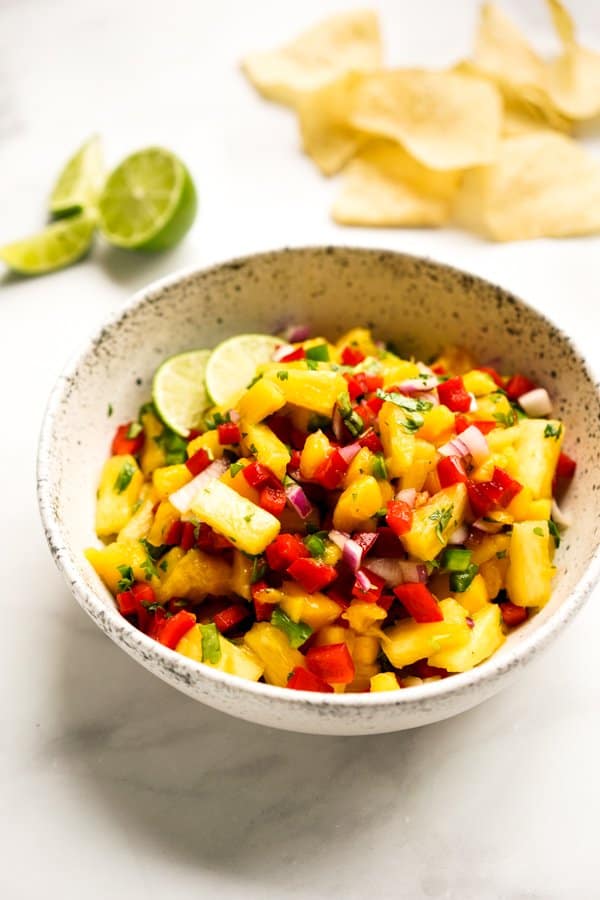 A bowl of pineapple chunks, mango chunks, bell pepper pieces with lime slices and tortilla chips in the background