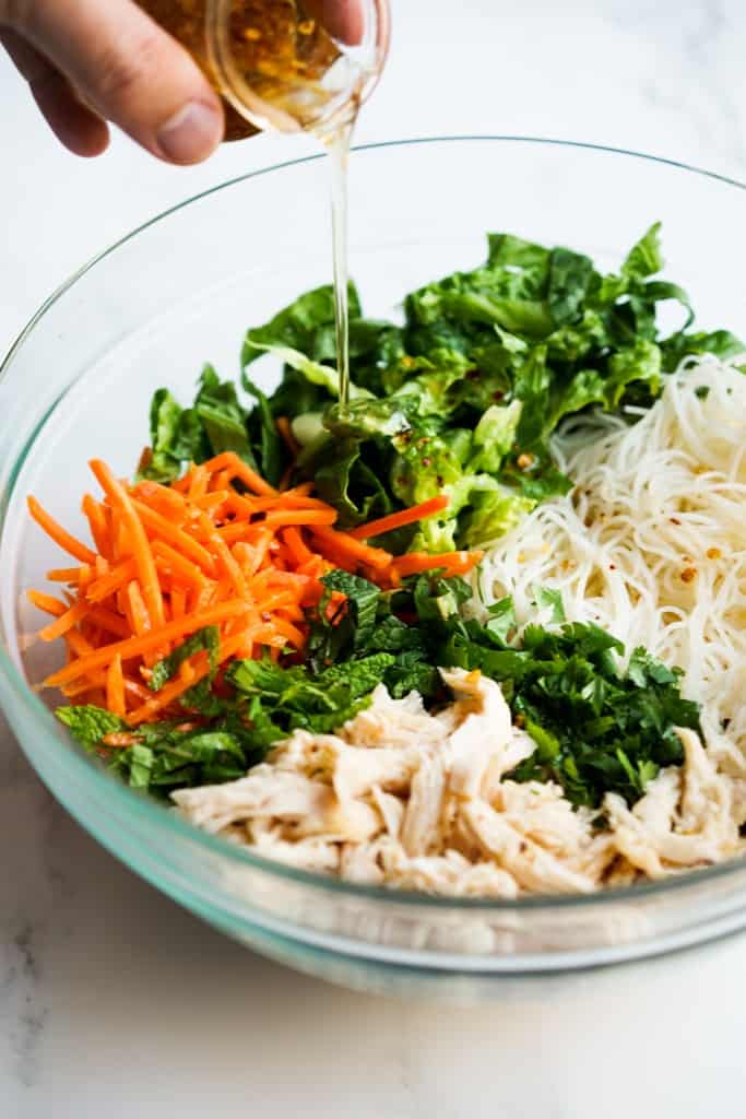 Drizzling dressing into a bowl of rice noodles, lettuce, carrots, cilantro, mint leaves and chicken