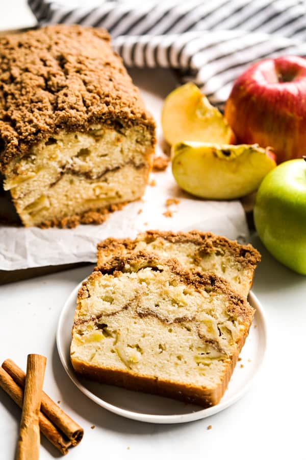 Two slices of Apple Cinnamon Bread on a plate and a loaf in the background, along with apples