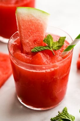 A glass of watermelon slushie with watermelons slices and mint leaves on it and around it