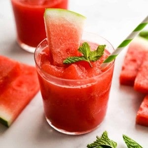 A glass of watermelon slushie with watermelons slices and mint leaves on it and around it