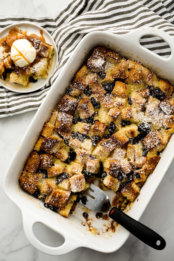 A small plate of old fashion bread pudding topped with vanilla ice cream next to the whole casserole dish of it