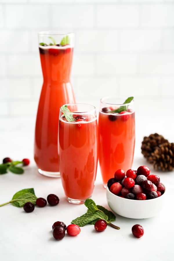 Two glasses of sparkling red beverage with a tall jug and a bowl of cranberries