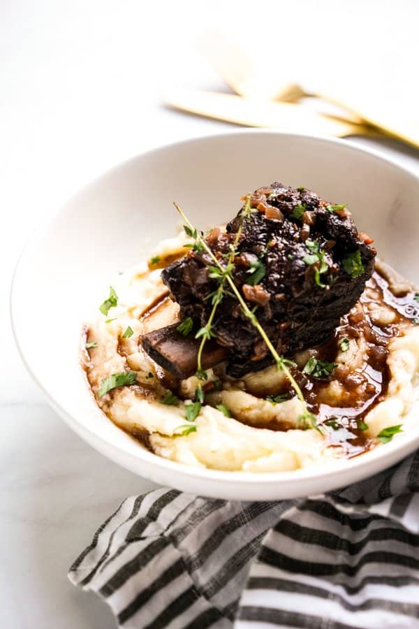 A bowl of mashed potatoes topped with a piece of beef short ribs, topped with herbs and gravy