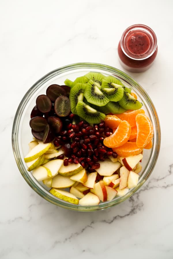 A large bowl consisting of kiwi, grapes, pears, apples, mandarin oranges and pomegranate. Dressing on the side 