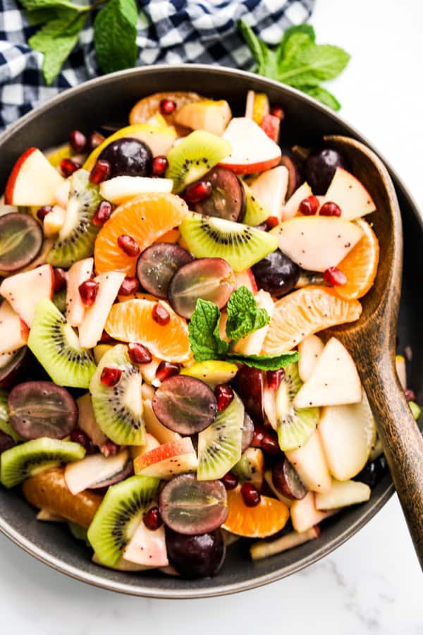 A brown ladle scooping out from a bowl of fruit salad with mandarin oranges, pear, apple, grapes kiwi and pomegranate