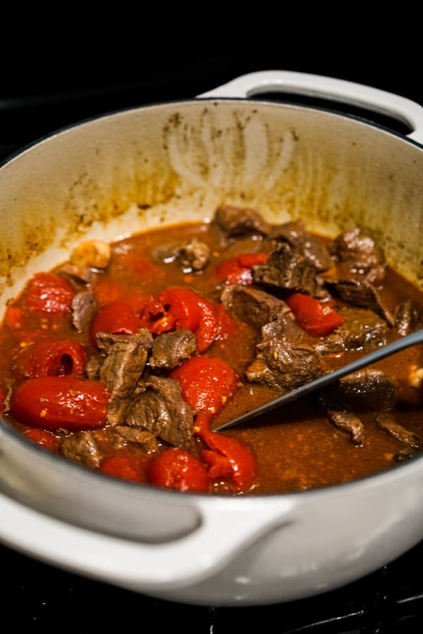 Breaking apart tomatoes in a pot also filled with beef chunks and broth