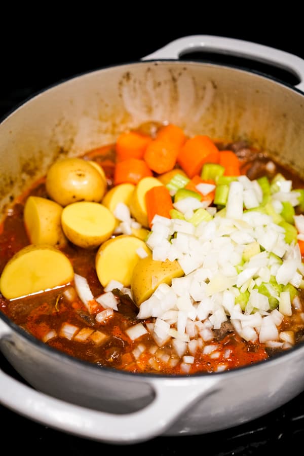 Adding potatoes, carrots, onions and celery to a pot of beef stew on the stove