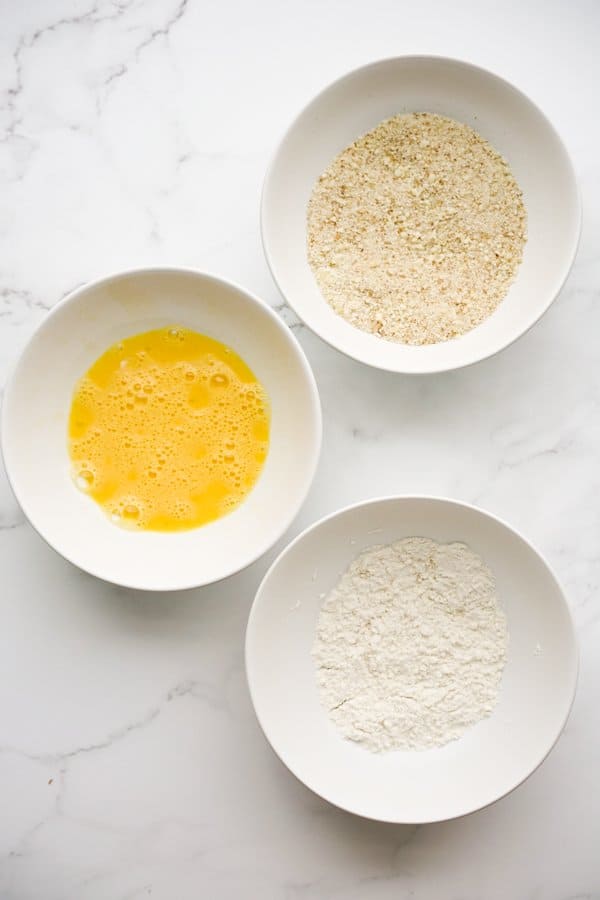 A bowl of flour, a bowl of whisked eggs, and a bowl of breadcrumbs and parmesan cheese