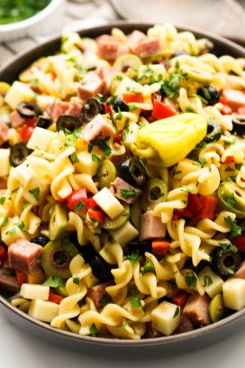 A bowl of pasta salad with ham, salami, olives and cubed cheeses