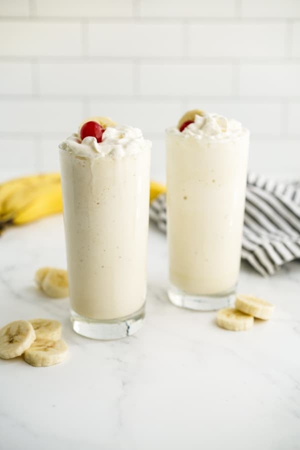 Two glasses of banana milkshake with whipped cream, banana and cherries on top