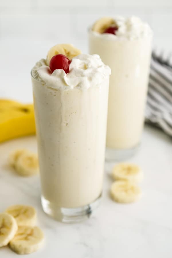 A closeup of a tall glass of Banana Milkshake topped with whipped cream, cherries and sliced banana