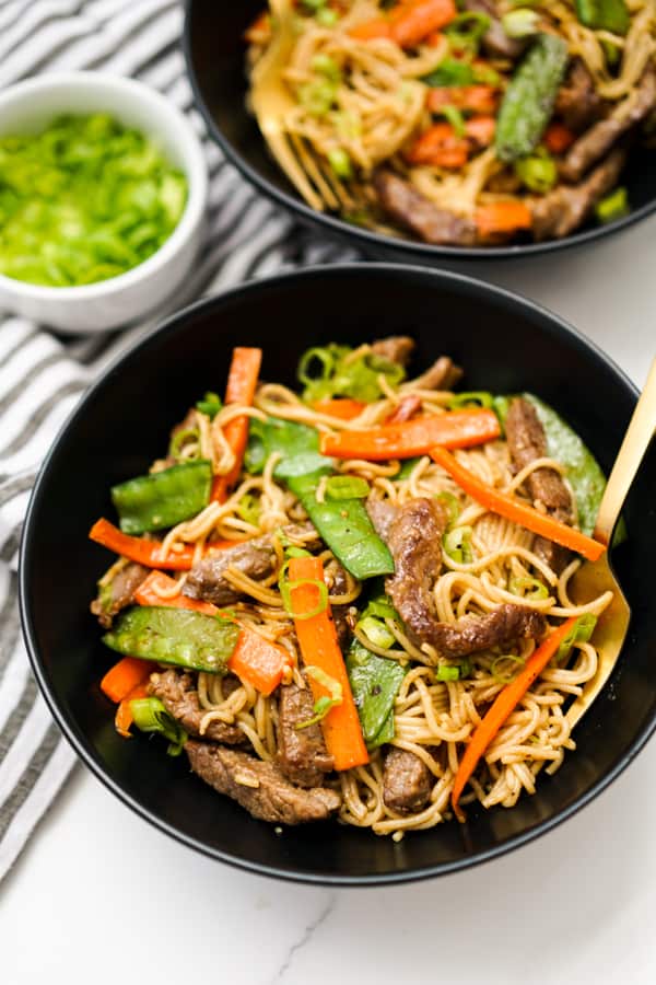 Two bowls of Mongolian Beef and noodles along with carrots and snap peas