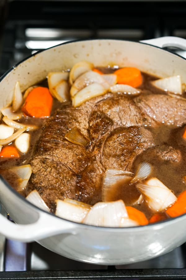 A large piece of beef chuck, carrots and onions in beef broth in Dutch oven on stovetop