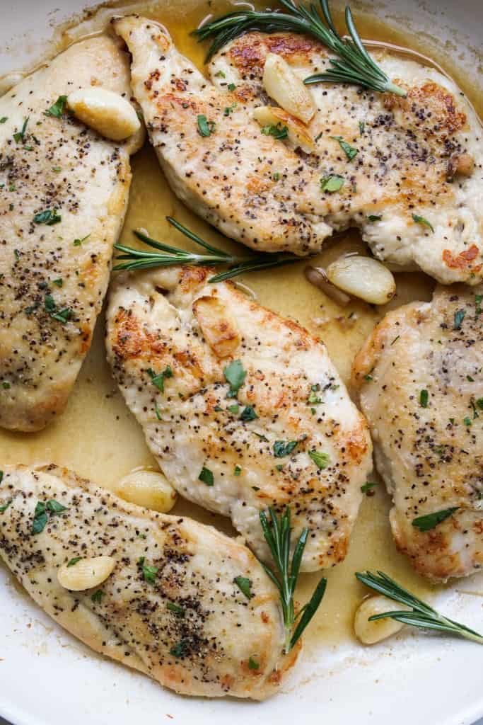 Skillet of chicken breasts sitting in sauce and surrounded with fresh rosemary twigs and garlic cloves