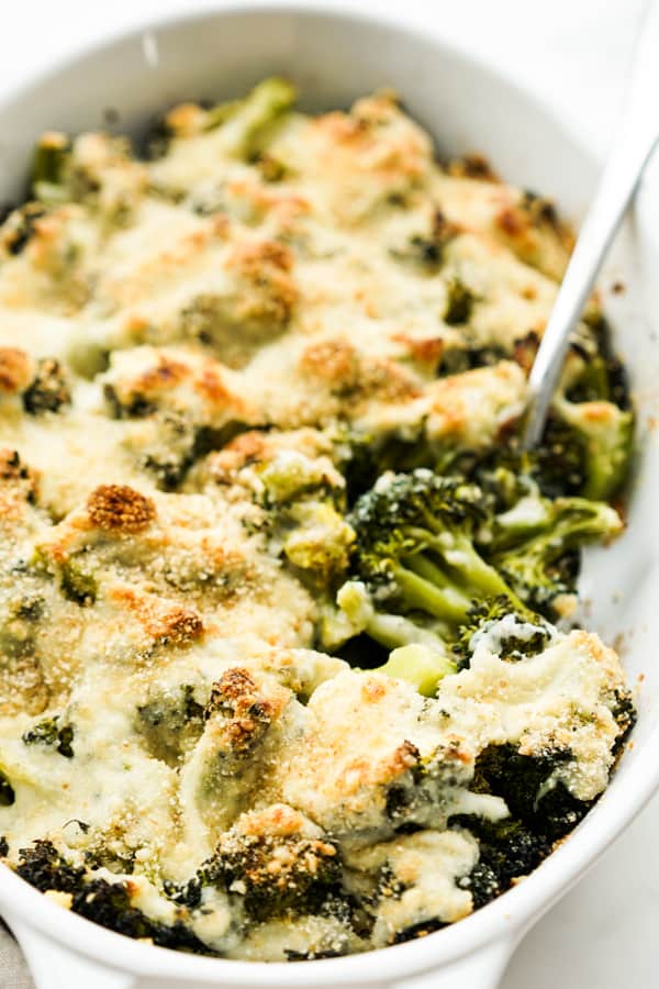 Spoon digging out broccoli from a oval casserole of broccoli au gratin