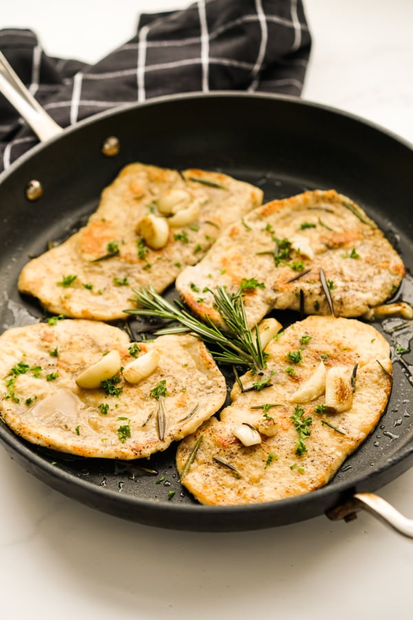 A skillet of thinly sliced chicken breast topped with garlic and rosemary