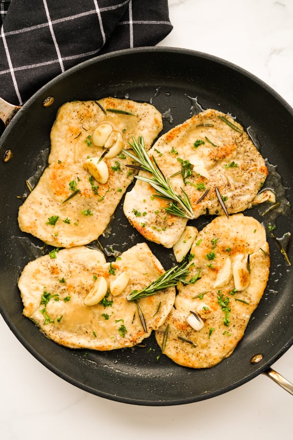 Top down view of a skillet with four pieces of Garlic Rosemary Chicken topped with herbs and garlic cloves
