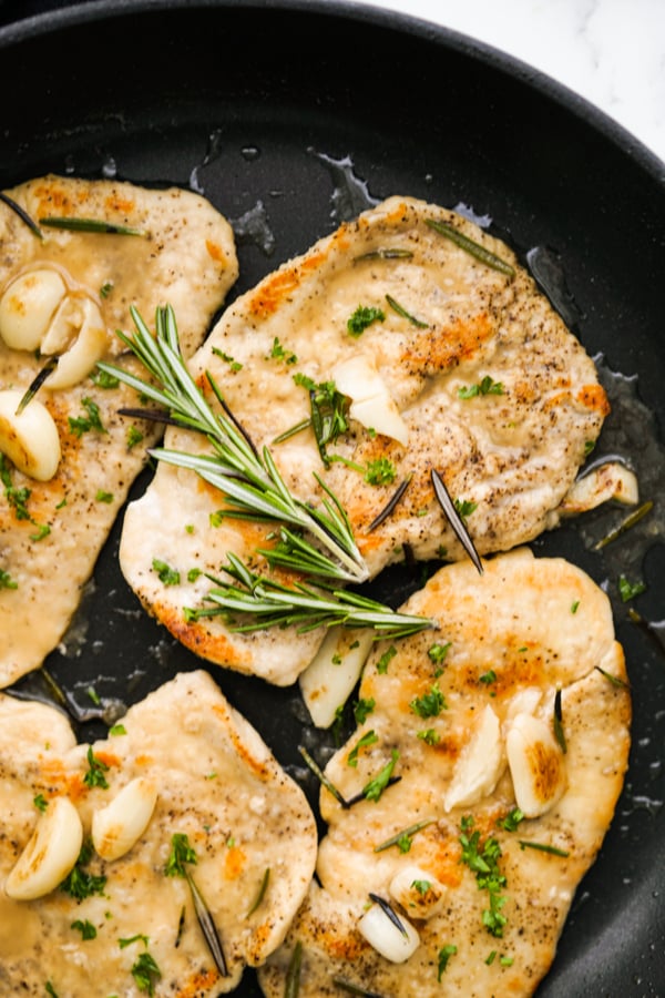 Chicken cutlets in a skillet topped with rosemary and garlic cloves