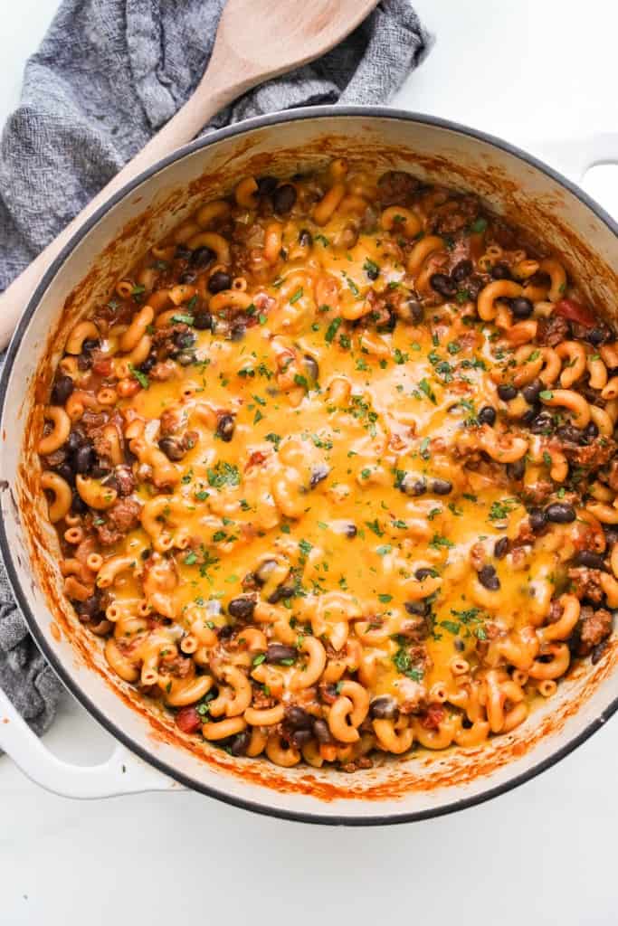 Top down view of a pot of chili macaroni and cheese topped with melted cheese, with a wooden spoon on the side