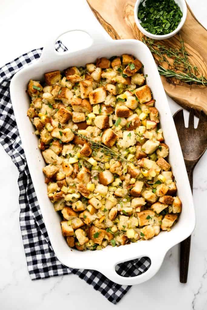 A rectangular casserole dish filled with apple stuffing, herbs on the side