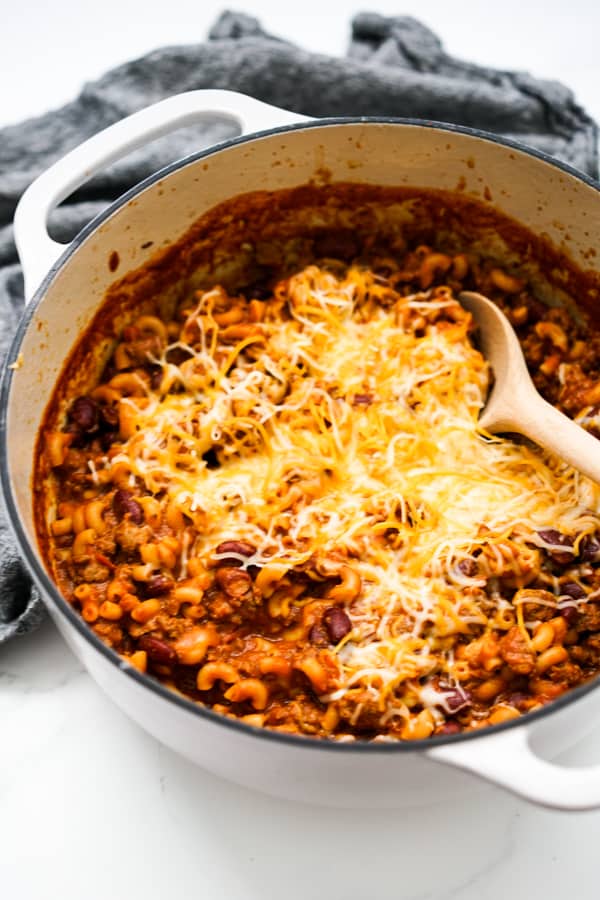 A large white pot of Chili Mac with shredded cheese on top and a wooden spoon