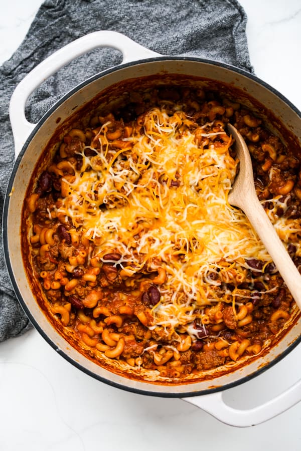 Chili mac and cheese topped with more cheese in a large pot with a wooden spoon