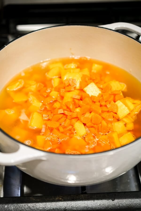 Chicken broth, butternut squash, and carrots cooking in a large white Dutch oven on the stove