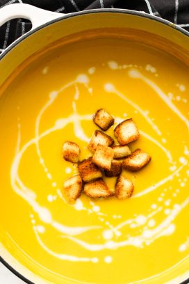 Top down view of a white pot of Curry Butternut Squash Soup with croutons and coconut swirls