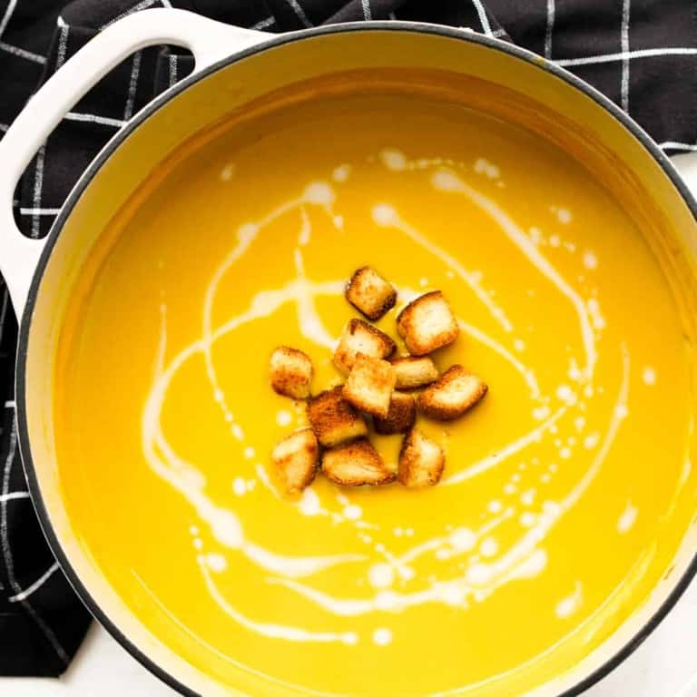 Top down view of a white pot of Curry Butternut Squash Soup with croutons and coconut swirls
