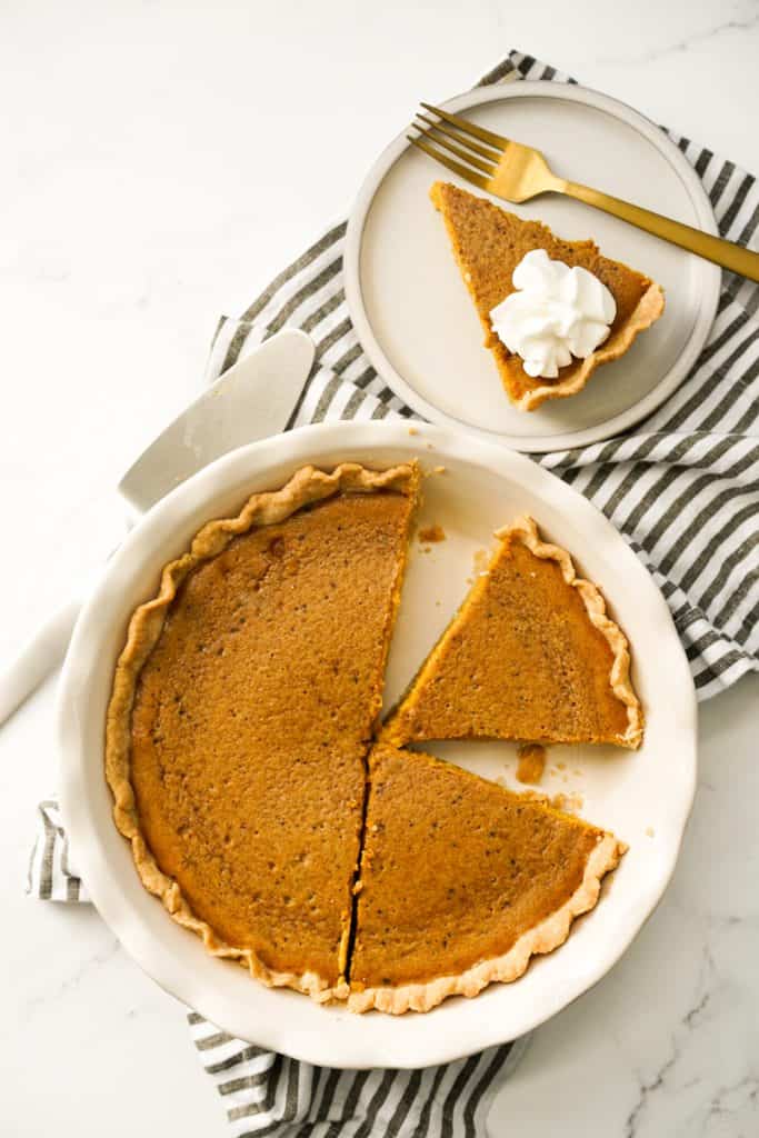 Top down view of a sliced whole pumpkin pie and a small plate of a slice with whipped cream on top