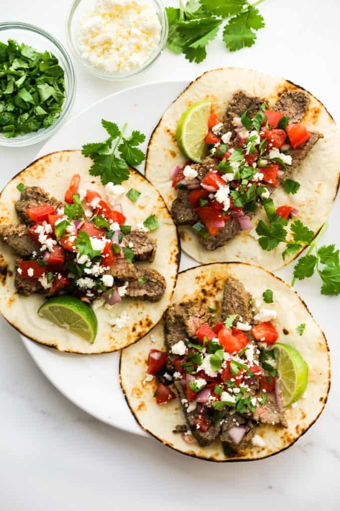 A plate of three grilled steak tacos with cheese and cilantro on the side.