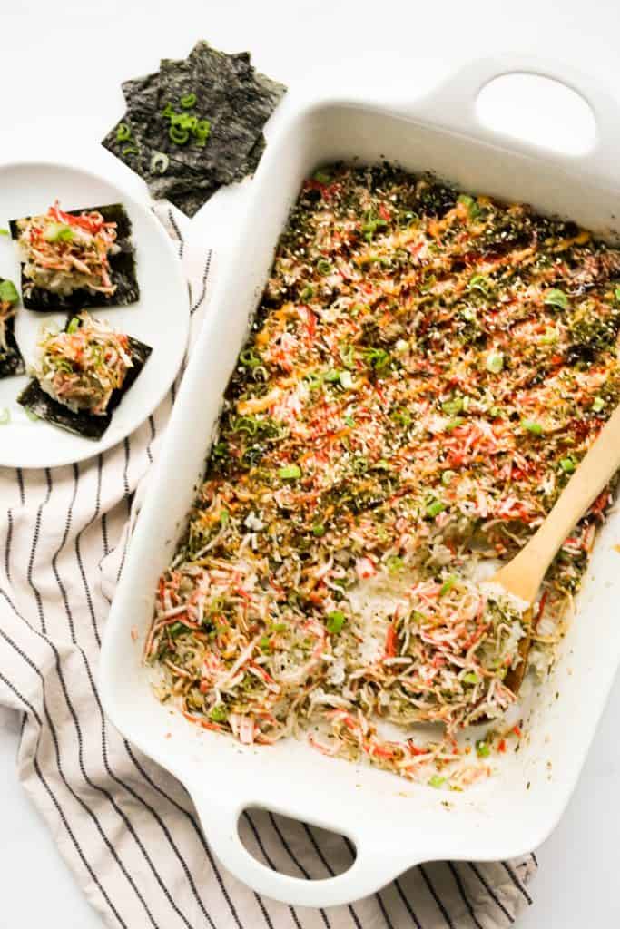 A spatula digging into a rectangular casserole of sushi bake, with small packets of rice wrapped in seaweed on the side