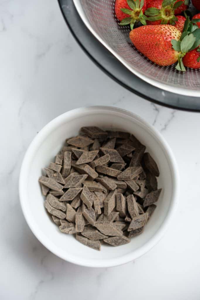 A bowl of chocolate chunks next to a colander of fresh strawberries