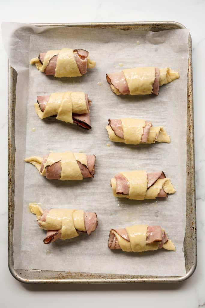 Unbaked crescent rolls rolled up with ham and cheese on a large baking sheet lined with parchment paper.