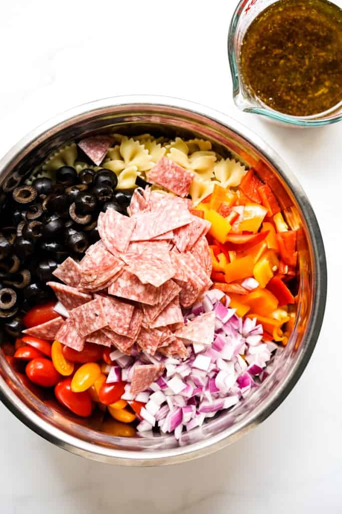 A mixing bowl filled with bow tie pasta, bell pepper, red onions, salami, black olives. Dressing on the side