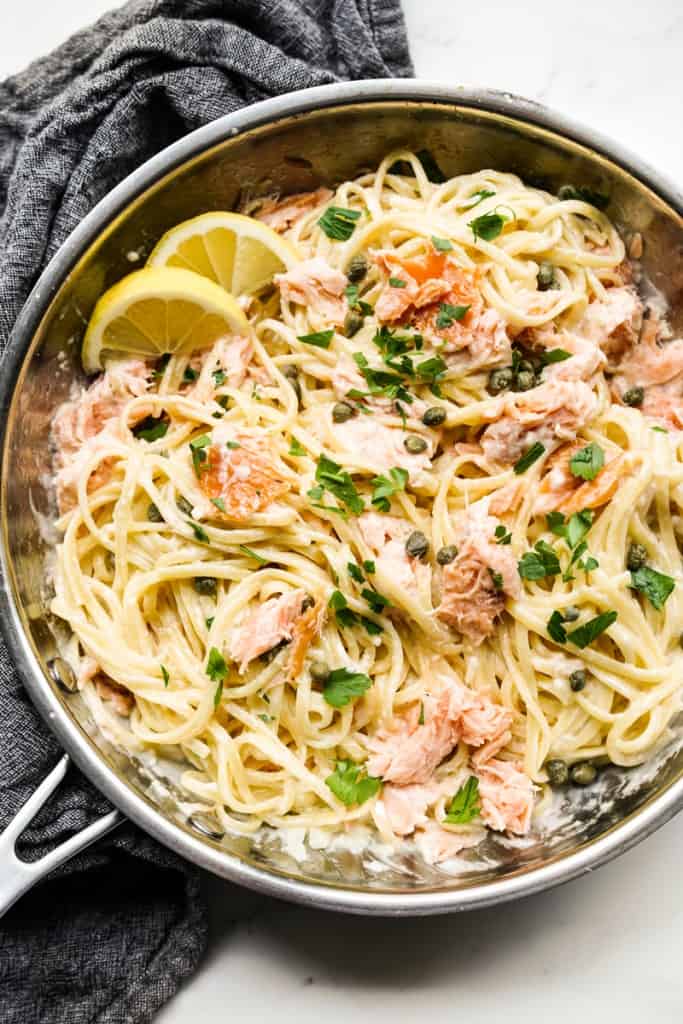A skillet filled with creamy linguini pasta tossed with smoked salmon and capers, with lemon wedges on the side
