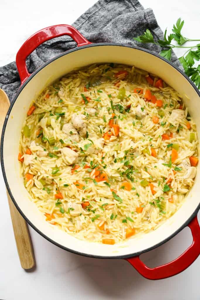 Top down view of a red Dutch oven filled with creamy chicken and orzo, along with carrots and celery