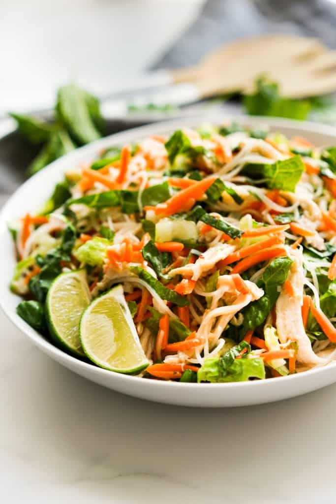 A bowl of Vietnamese noodle salad loaded with shredded carrots, lettuce, mint leaves, cilantro, chicken and wedges of lime