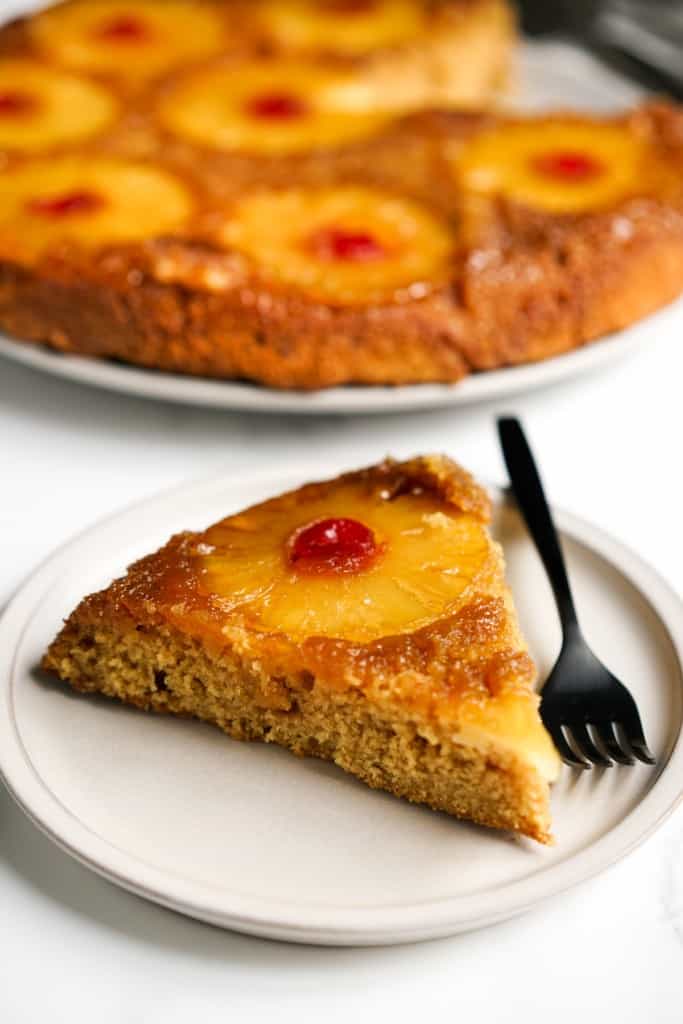 A slice of skillet pineapple upside down cake on small plate, with the large cake in the background