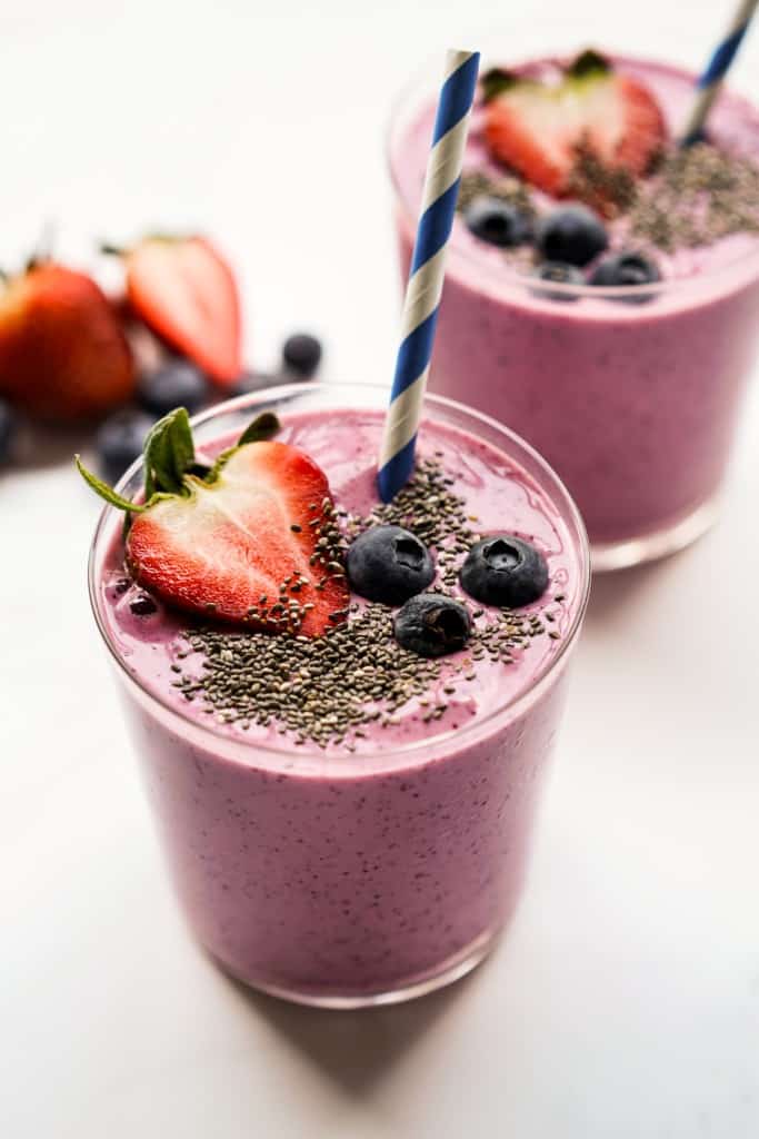 Two glasses of pink colored thick smoothie topped with strawberries, blueberries and bananas, with blue stripes straws